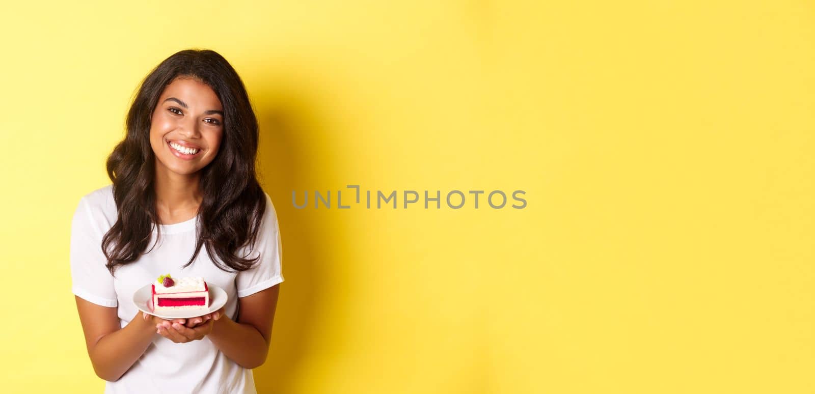 Portrait of cute african-american girl, holding piece of cake and smiling, standing over yellow background.