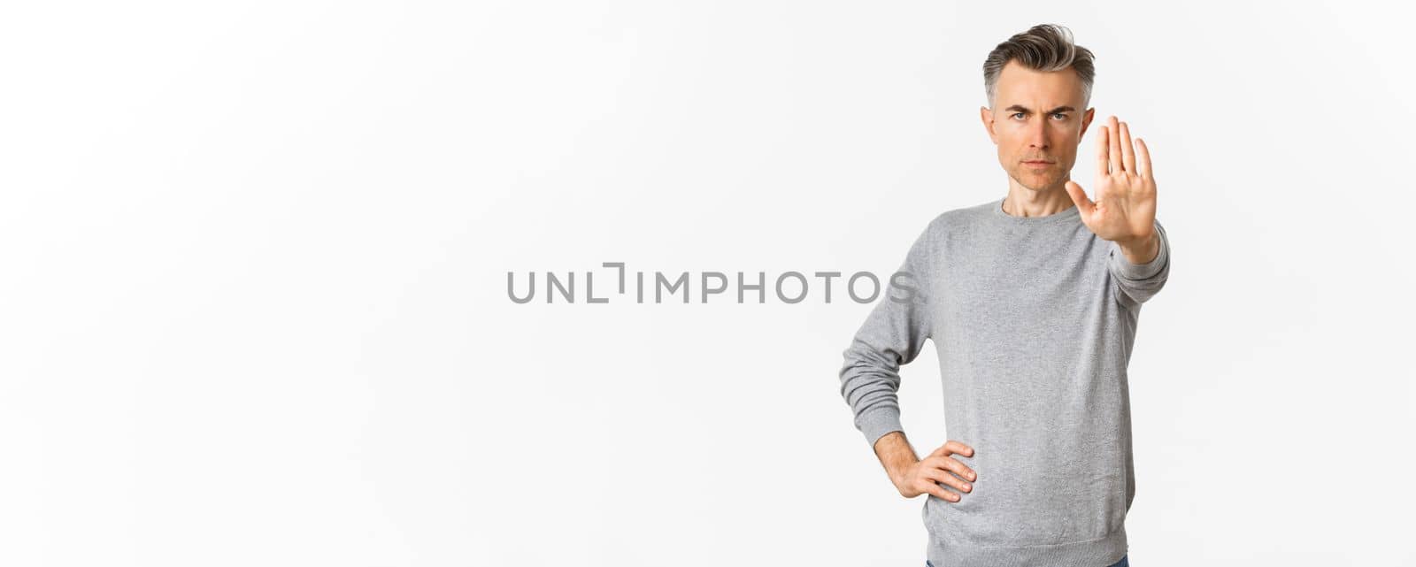 Image of serious middle-aged man extend one hand to stop you, prohibit something bad, frowning and looking with disapproval, standing over white background.