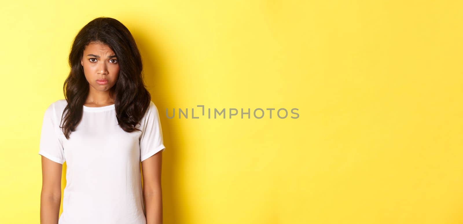 Portrait of gloomy and sad african-american girl, sulking and looking upset, standing in white t-shirt against yellow background.