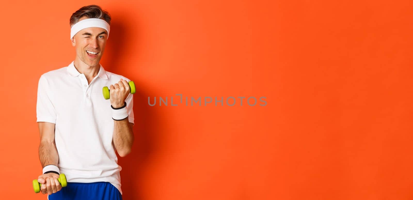 Portrait of handsome cheeky, middle-aged male athlete doing sports, exercise with dumbbells and winking at camera, standing over orange background by Benzoix