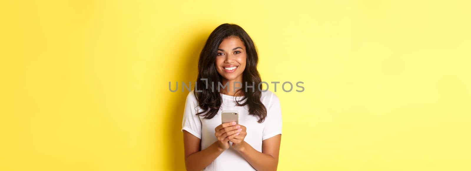 Image of modern african-american girl smiling, using mobile phone, standing over yellow background.