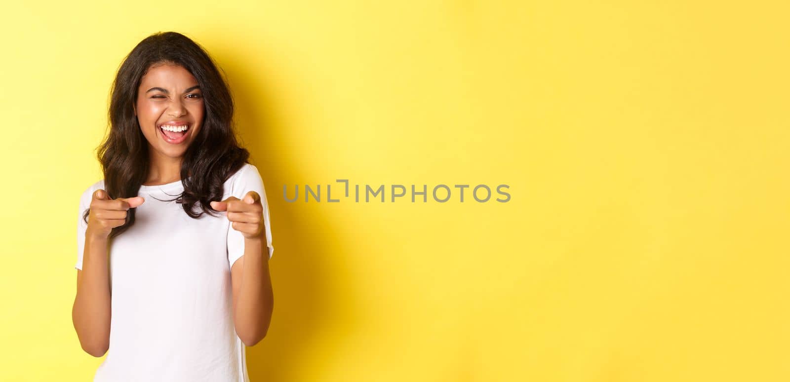 Portrait of cheeky, beautiful african-american woman in white t-shirt, winking and pointing fingers at camera to say congrats, praising you, standing over yellow background by Benzoix