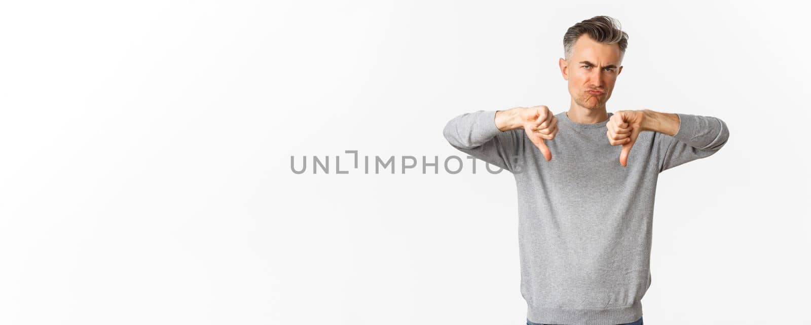 Skeptical and disappointed middle-aged man, grimacing unamused and showing thumbs-down, dislike something bad, standing over white background.