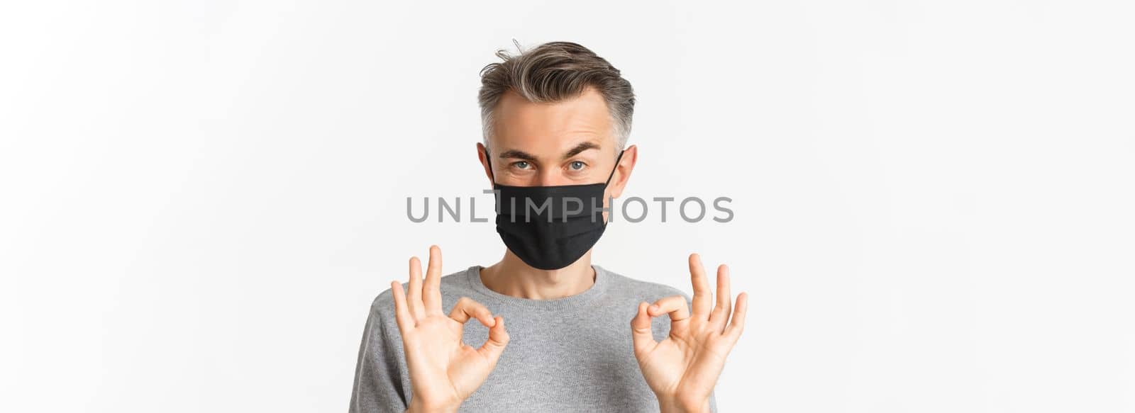 Concept of covid-19, social distancing and quarantine. Close-up of confident middle-aged man, wearing black medical mask, showing okay signs and praising something good.