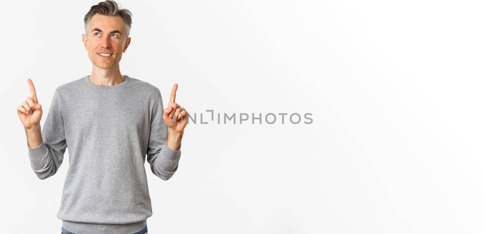 Image of handsome middle-aged man in grey sweater making his choice, smiling thoughtful, pointing fingers up and looking at product, standing over white background.