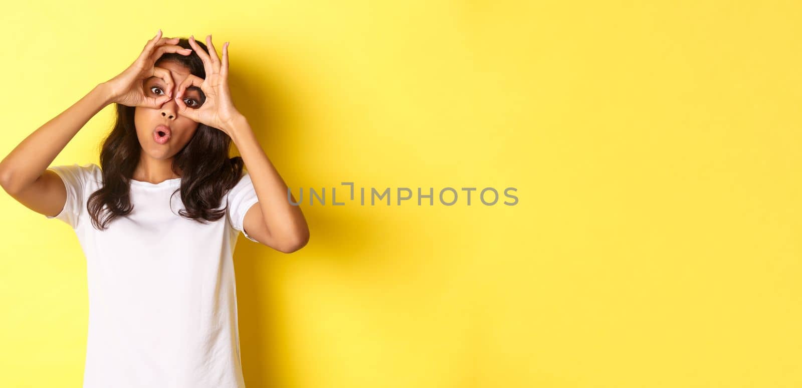 Image of happy and funny african-american girl, making finger glasses and looking through them at something amazing, standing over yellow background.