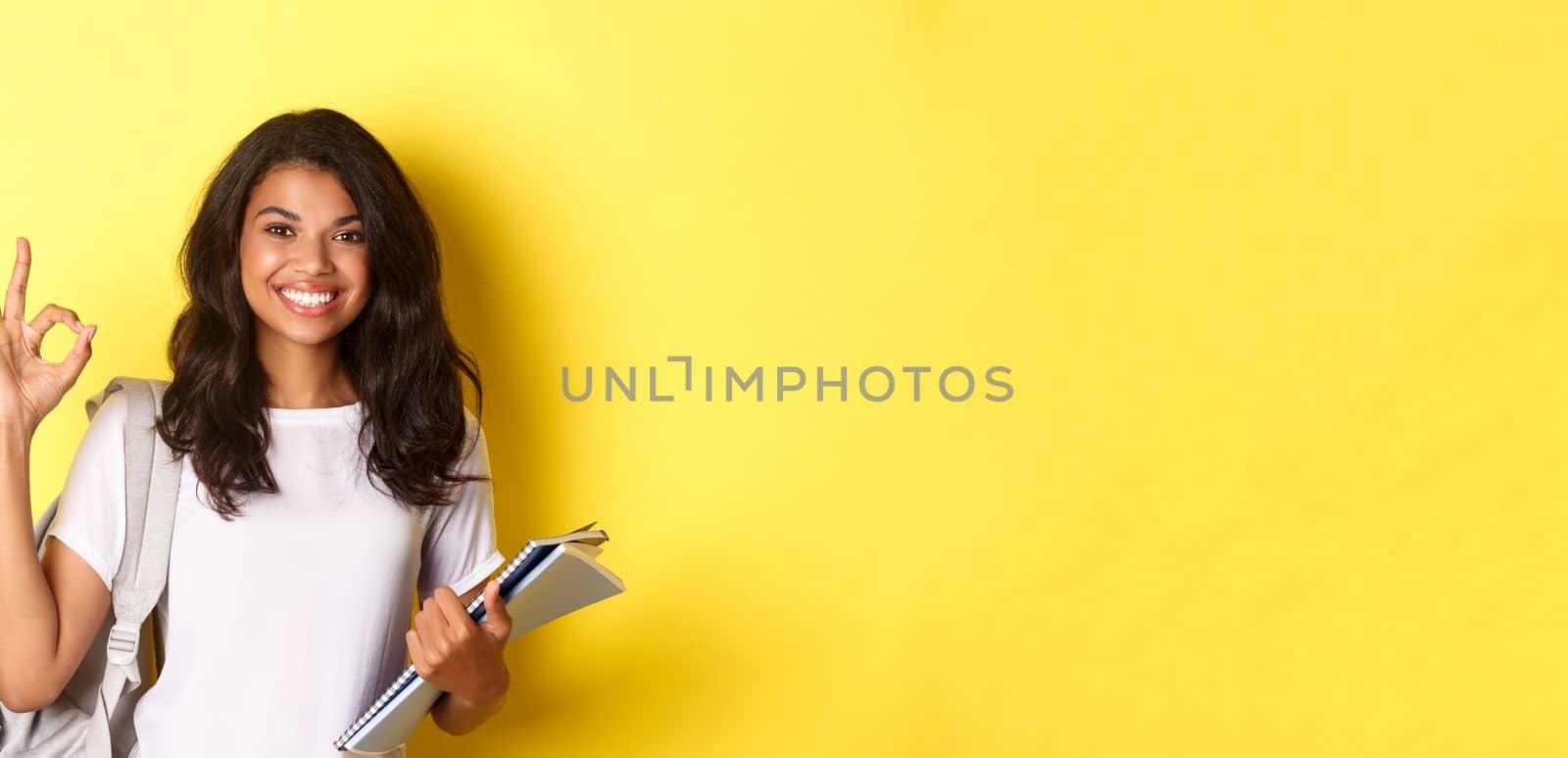 Portrait of satisfied african-american female student, smiling pleased and showing okay sign, like something good, standing over yellow background.