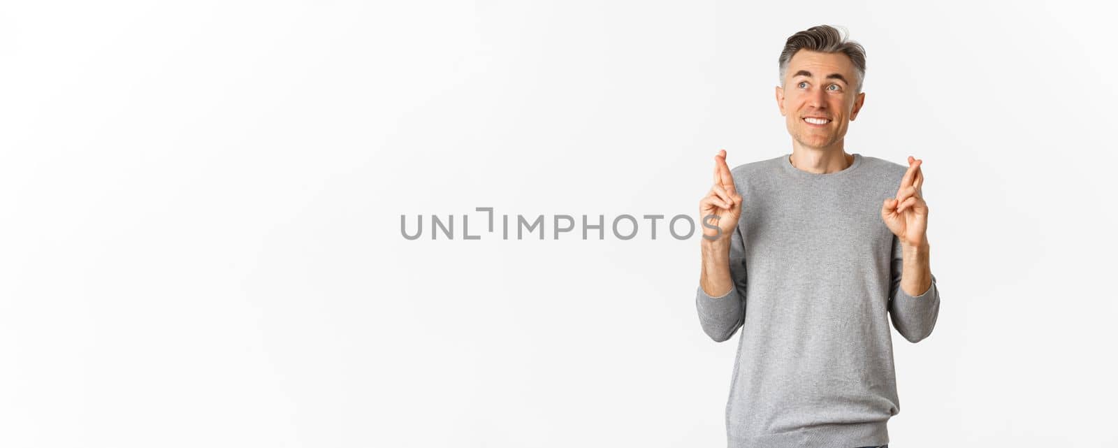 Image of excited and hopeful middle-aged man making a wish, waiting for results, cross fingers for good luck and looking at upper left corner, standing over white background.