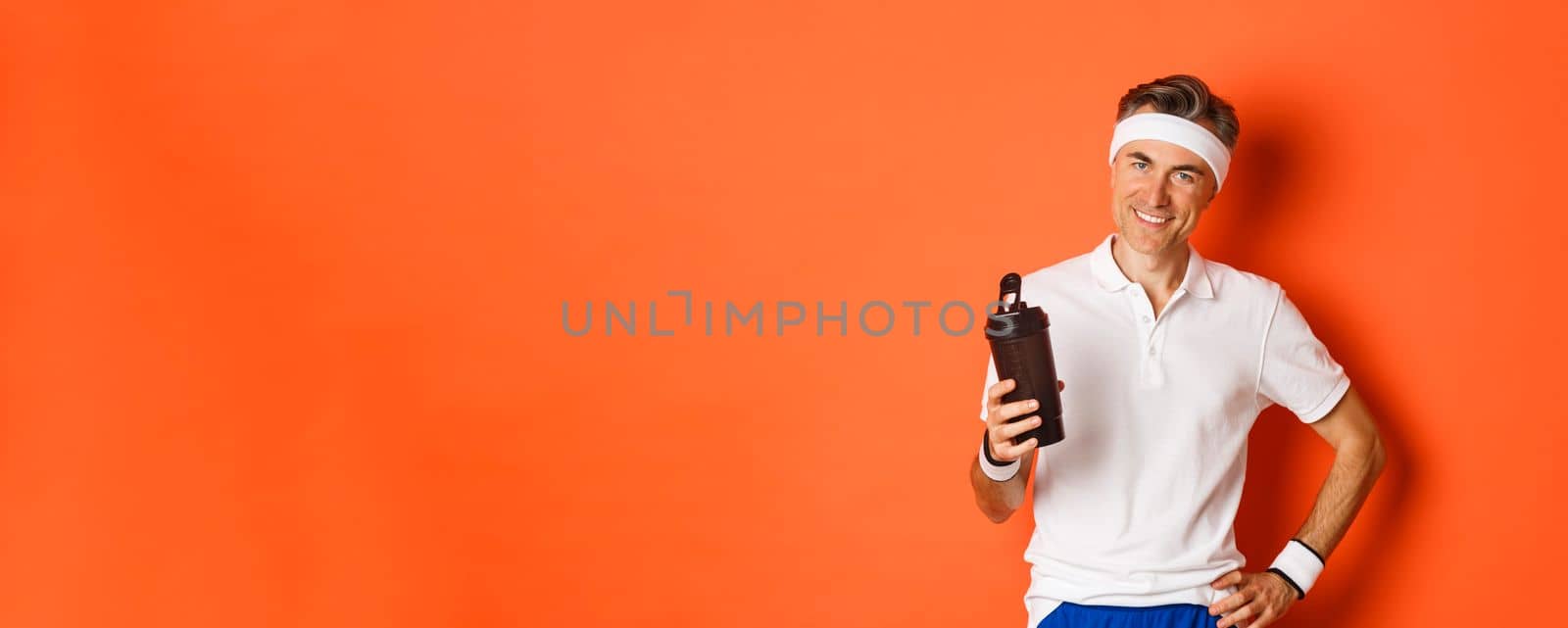 Portrait of healthy and fit male athlete, wearing gym uniform, drinking water or protein and smiling pleased, standing over orange background.