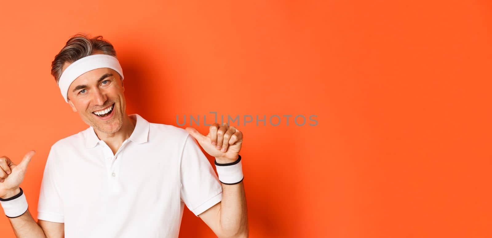 Close-up of confident middle-aged male athlete, feeling pleased, pointing at himself proudly, achieve goal, standing over orange background.