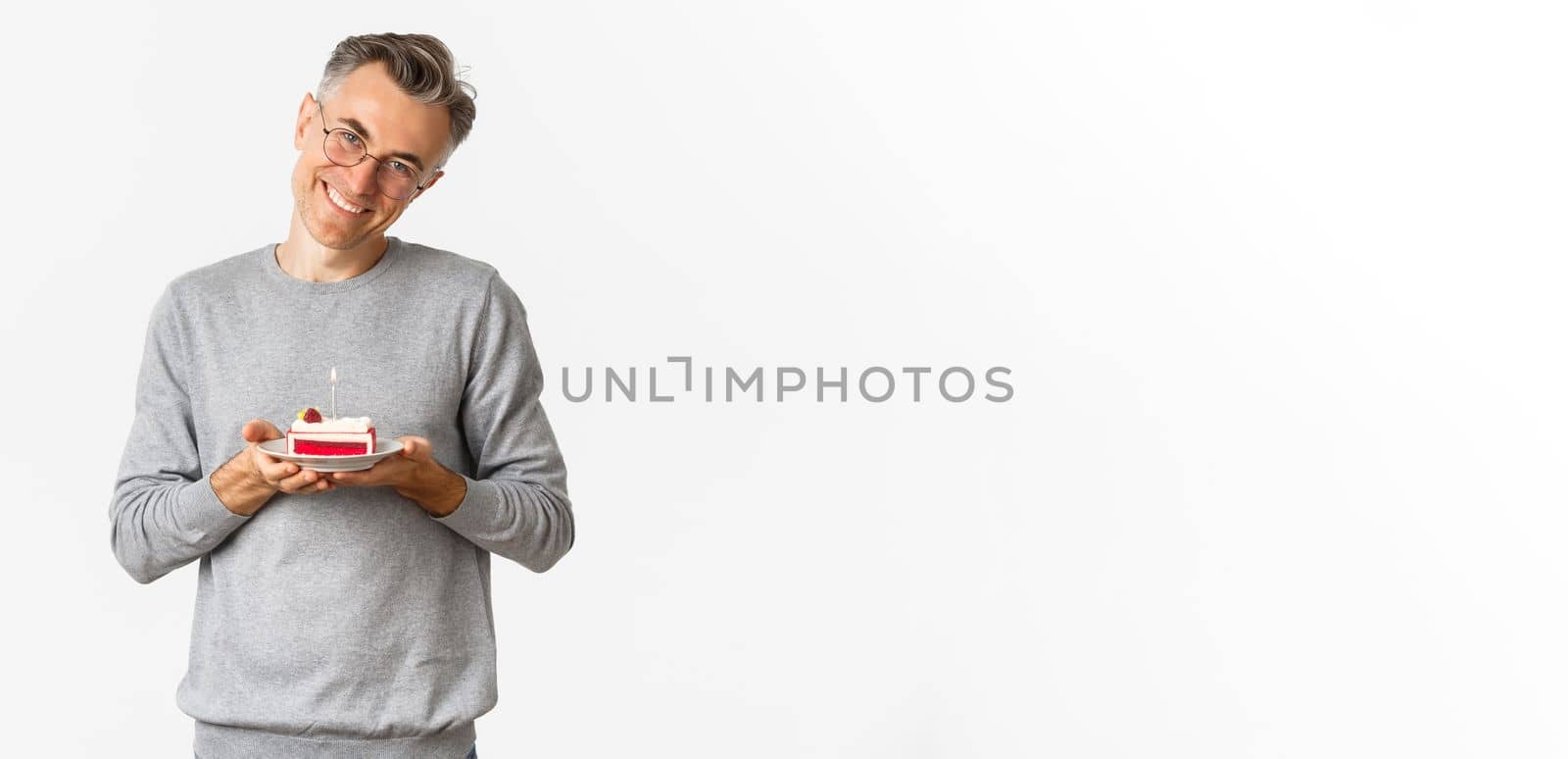 Portrait of smiling middle-aged man, looking touched and happy, celebrating birthday, holding b-day cake and celebrating, standing over white background.