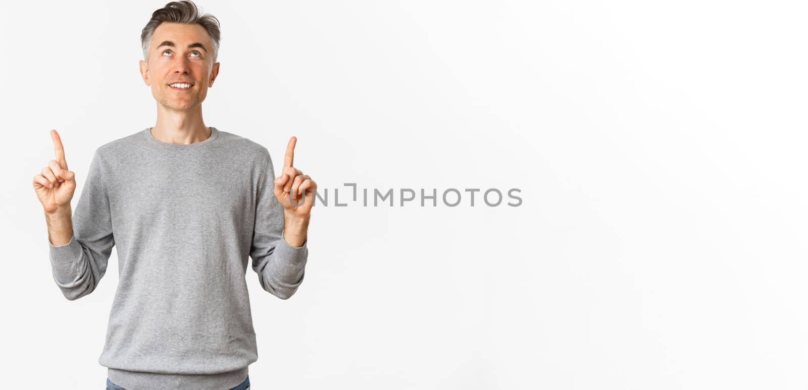 Portrait of smiling handsome man with short hair hair, looking and pointing fingers up at something interesting, reading an advertisement, standing over white background.