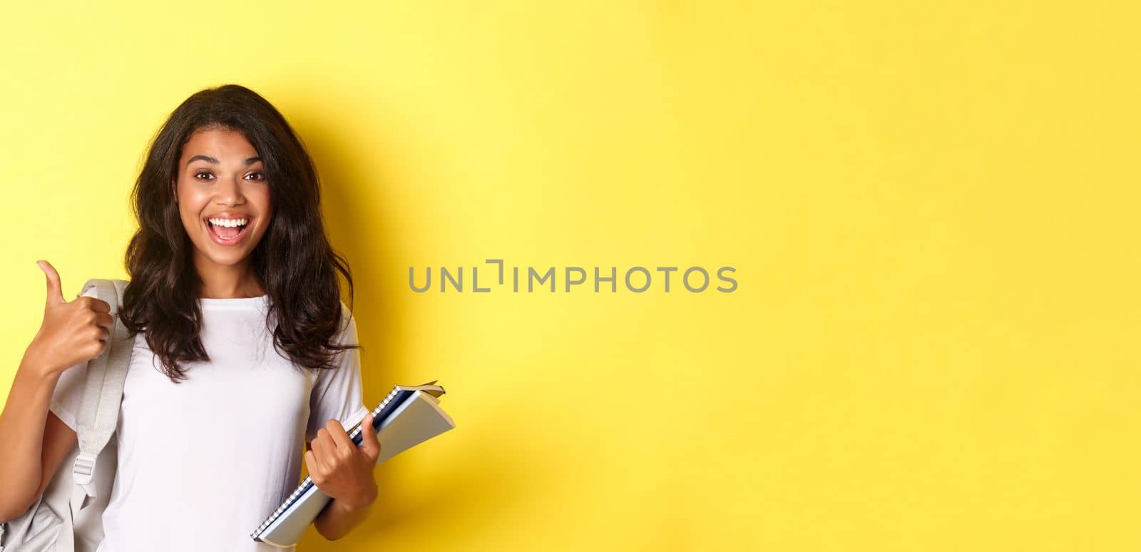 Image of cheerful african-american girl student, holding notebooks for classes and backpack, showing thumbs-up, like studying, standing over yellow background by Benzoix