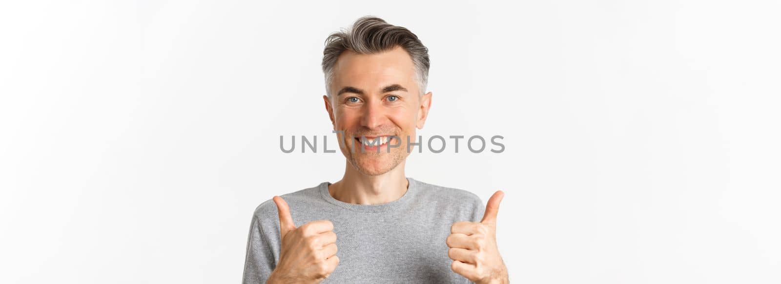 Close-up of cheerful middle-aged man, smiling happy and satisfied, showing thumbs-up, expressing approval, like something good, standing over white background.
