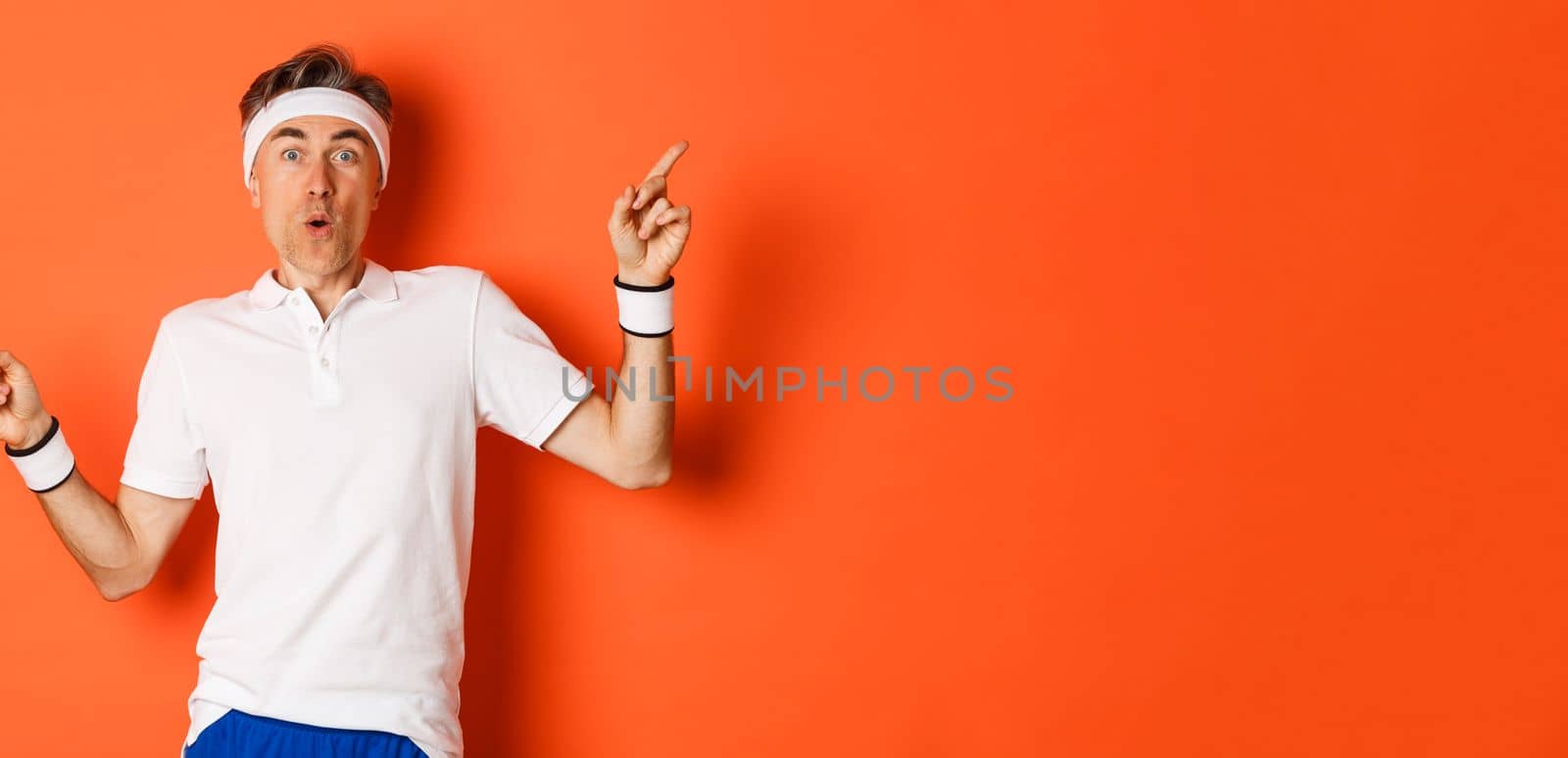 Concept of sport, fitness and lifestyle. Portrait of amazed and excited middle-aged male athlete, pointing fingers sideways, showing two variants, standing over orange background.