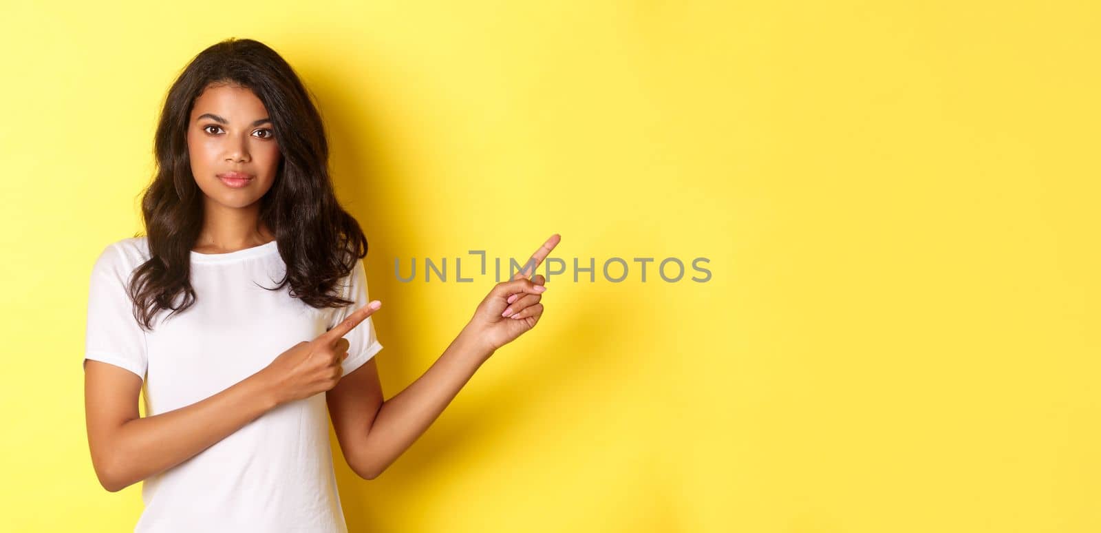 Image of confident and attractive african-american woman, pointing fingers right and looking at you assertive, standing over yellow background.