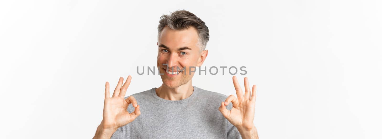 Close-up of handsome middle-aged man, smiling and showing okay signs, approve something or recommending, standing over white background.