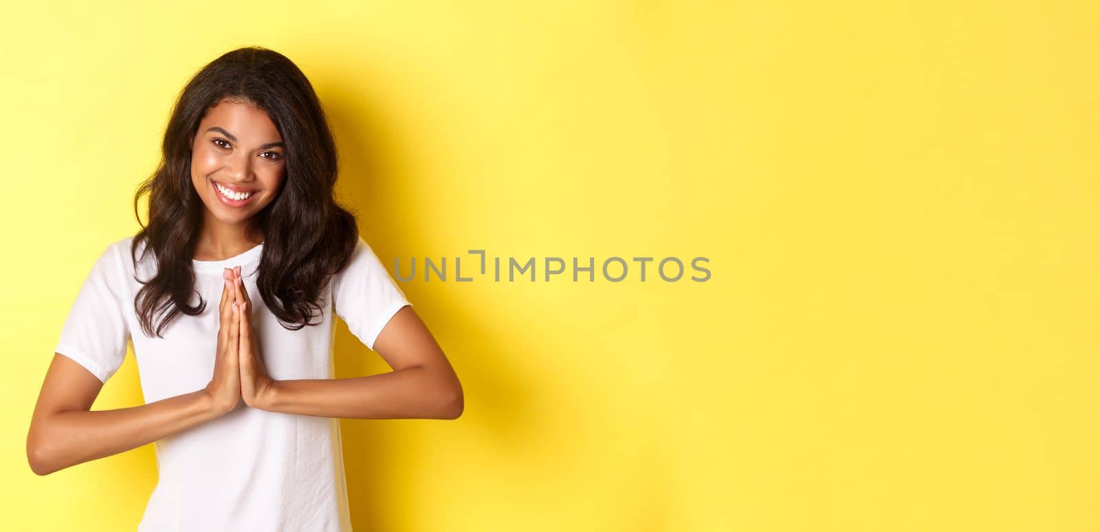 Image of cute african american woman, saying thank you and smiling, press hands together to express gratitude, standing over yellow background by Benzoix