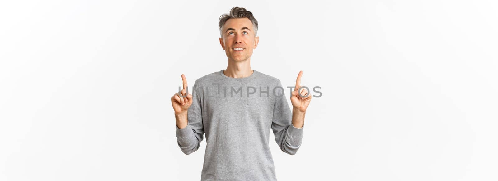 Portrait of smiling handsome man with short hair hair, looking and pointing fingers up at something interesting, reading an advertisement, standing over white background.