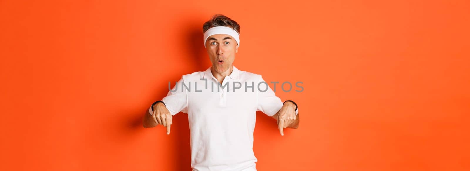 Concept of workout, sports and lifestyle. Portrait of surprised middle-aged male athlete, wearing clothes for gym, pointing fingers down and looking amazed, standing over orange background.