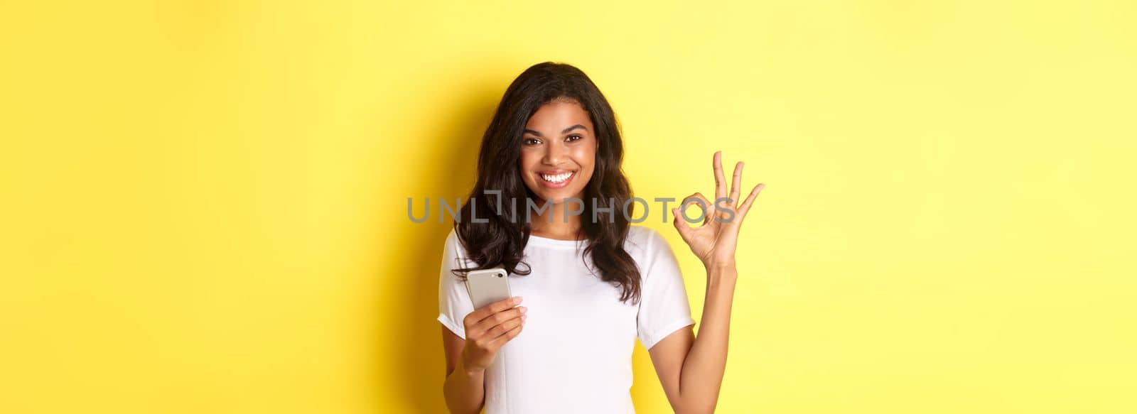 Image of good-looking african-american woman, showing okay sign with confidence, holding smartphone, standing over yellow background.