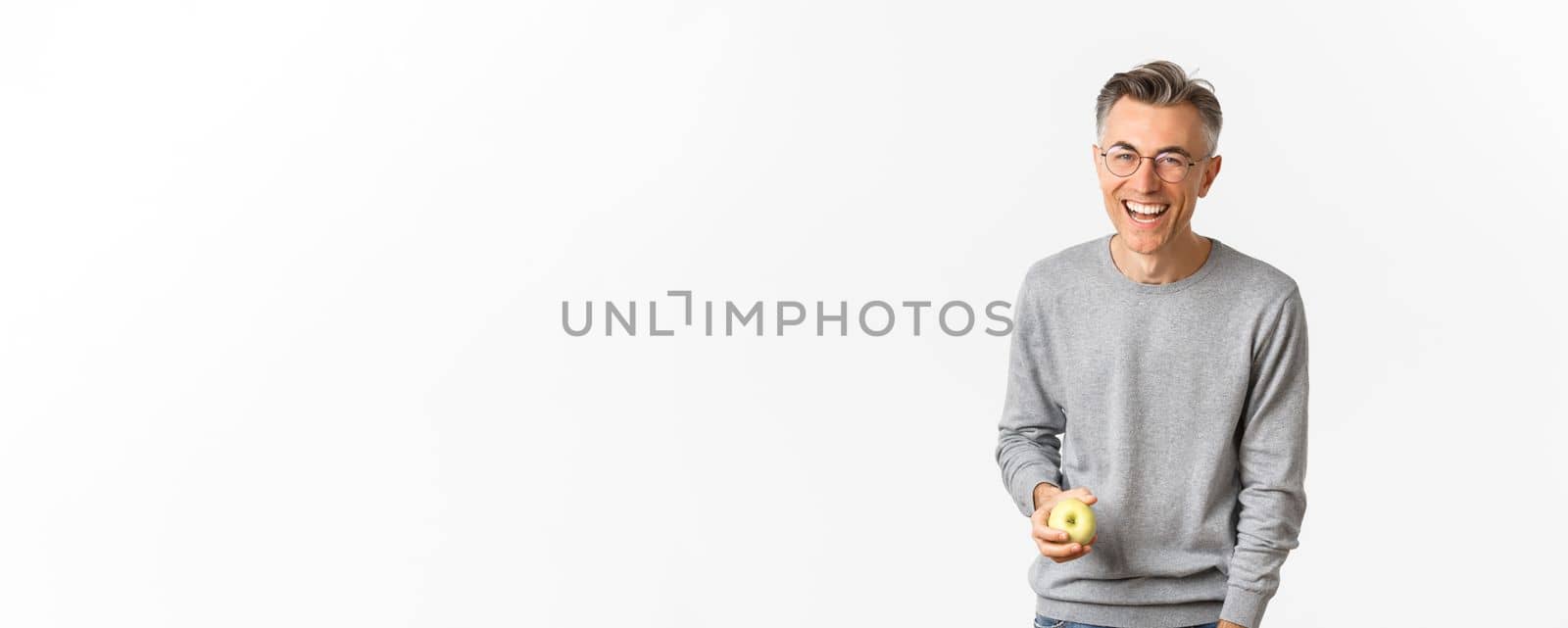 Image of happy and healthy middle-aged man, holding green apple, laughing and smiling carefree, standing over white background.