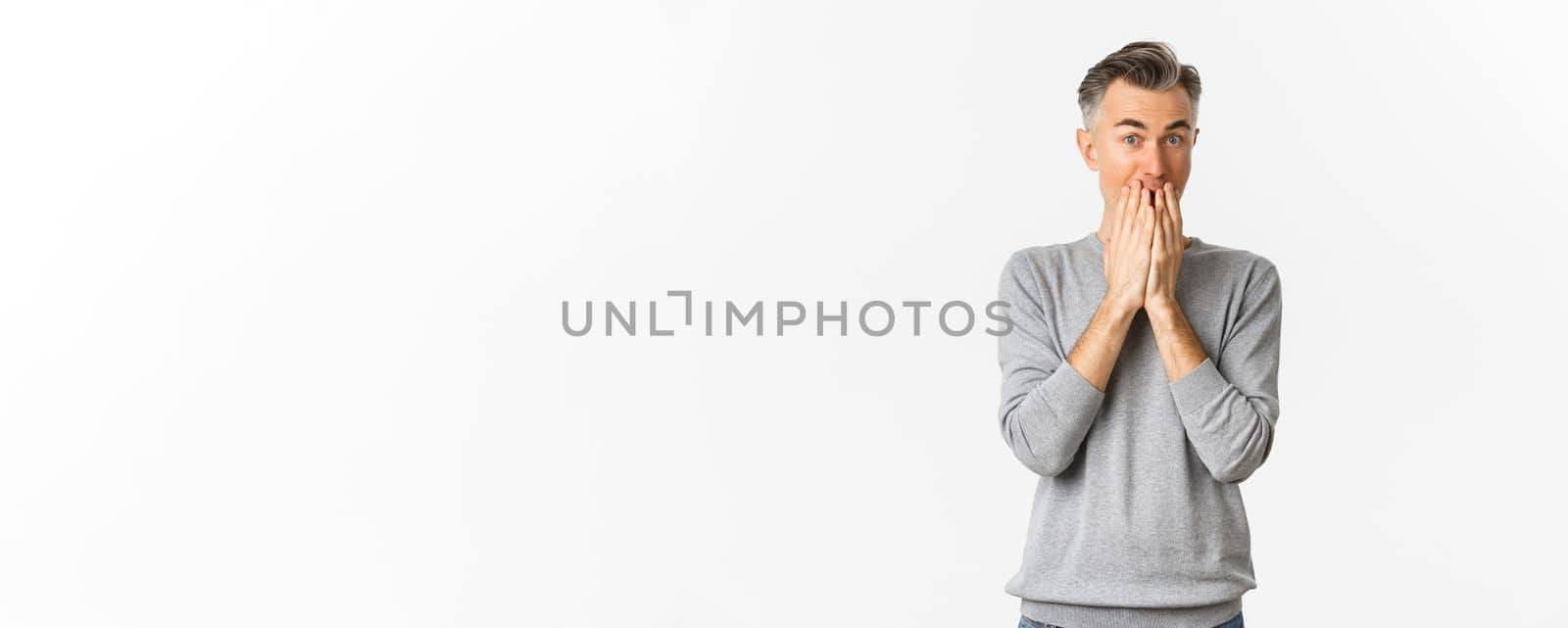 Image of excited and amused caucasian man with gray hair, cover mouth and looking at something interesting, standing over white background.