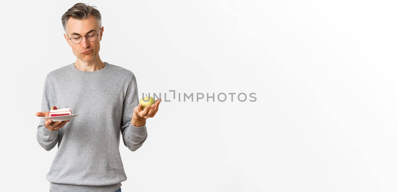 Portrait of thoughtful middle-aged man in glasses, making decision between tasty cake and green apple, standing over white background.