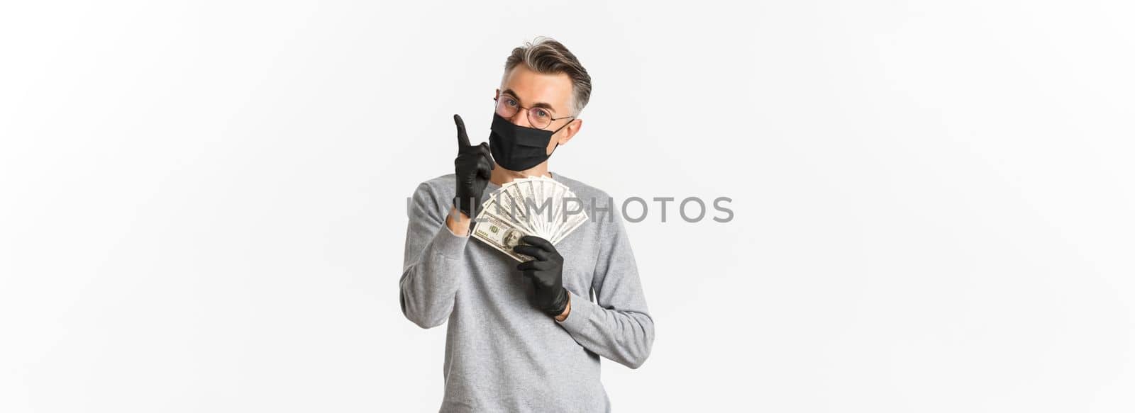 Concept of covid-19, social distancing and lifestyle. Portrait of lucky middle-aged man in medical mask, gloves and glasses, showing money and pointing finger up, smiling pleased.