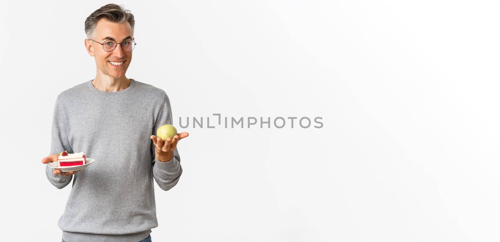 Image of handsome and healthy middle-aged man in glasses, recommending eat apple instead of cake, standing over white background.