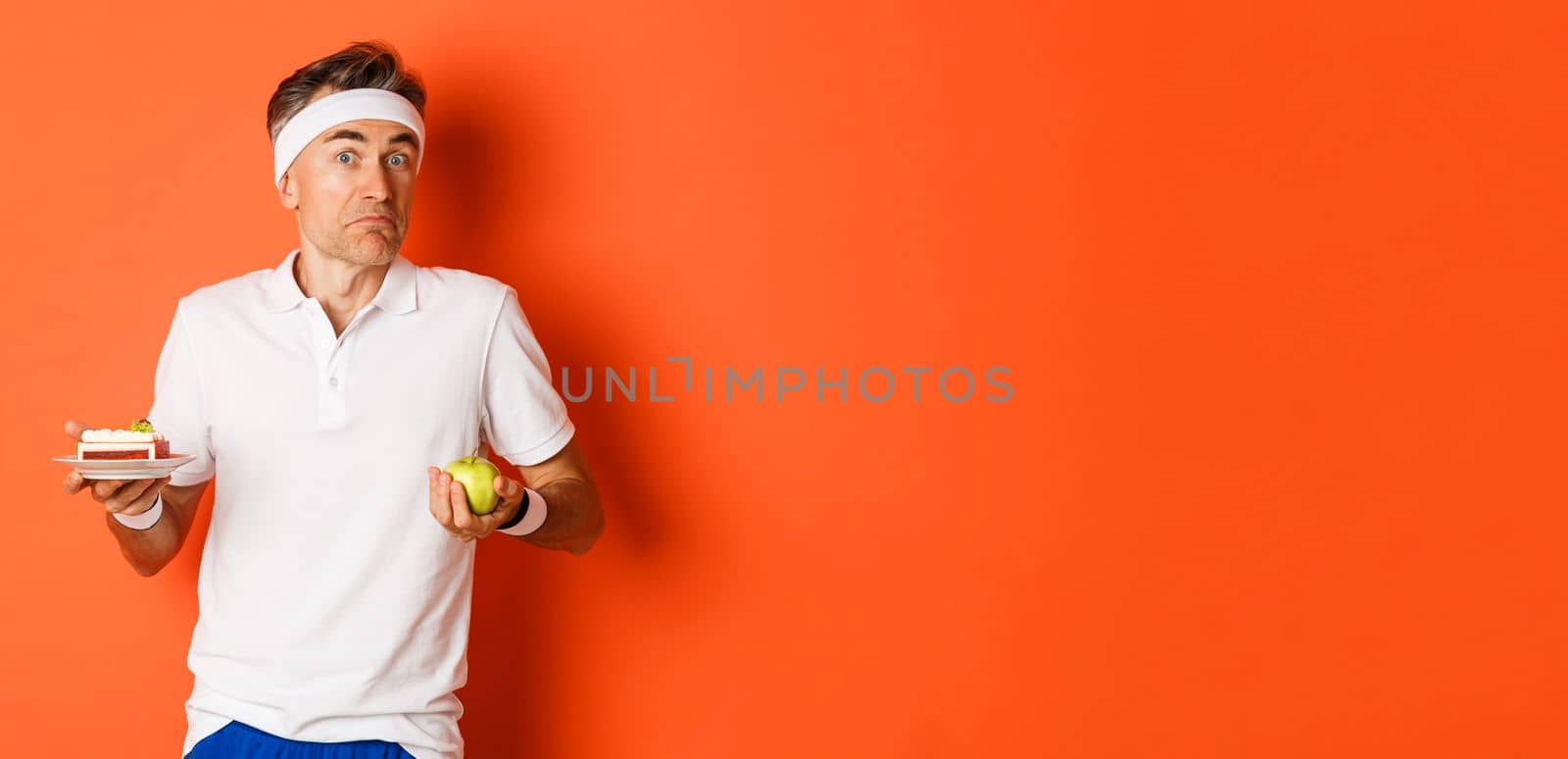 Image of clueless middle-aged fitness guy, holding apple and cake, shrugging shoulders, standing in workout uniform over orange background.