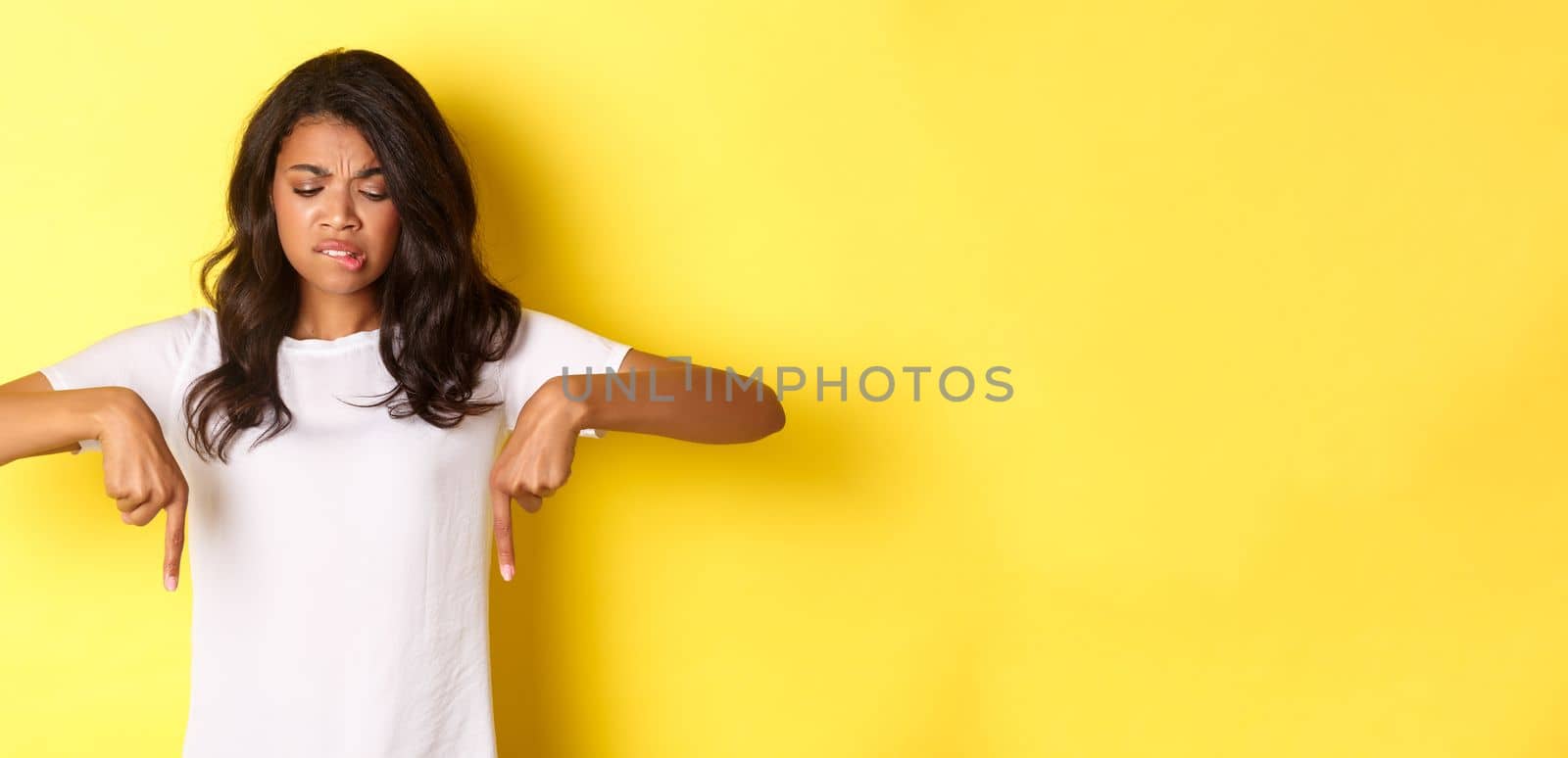 Portrait of doubtful and skeptical african-american girl in white t-shirt, frowning and pointing fingers down at something strange of unpleasant, standing over yellow background.