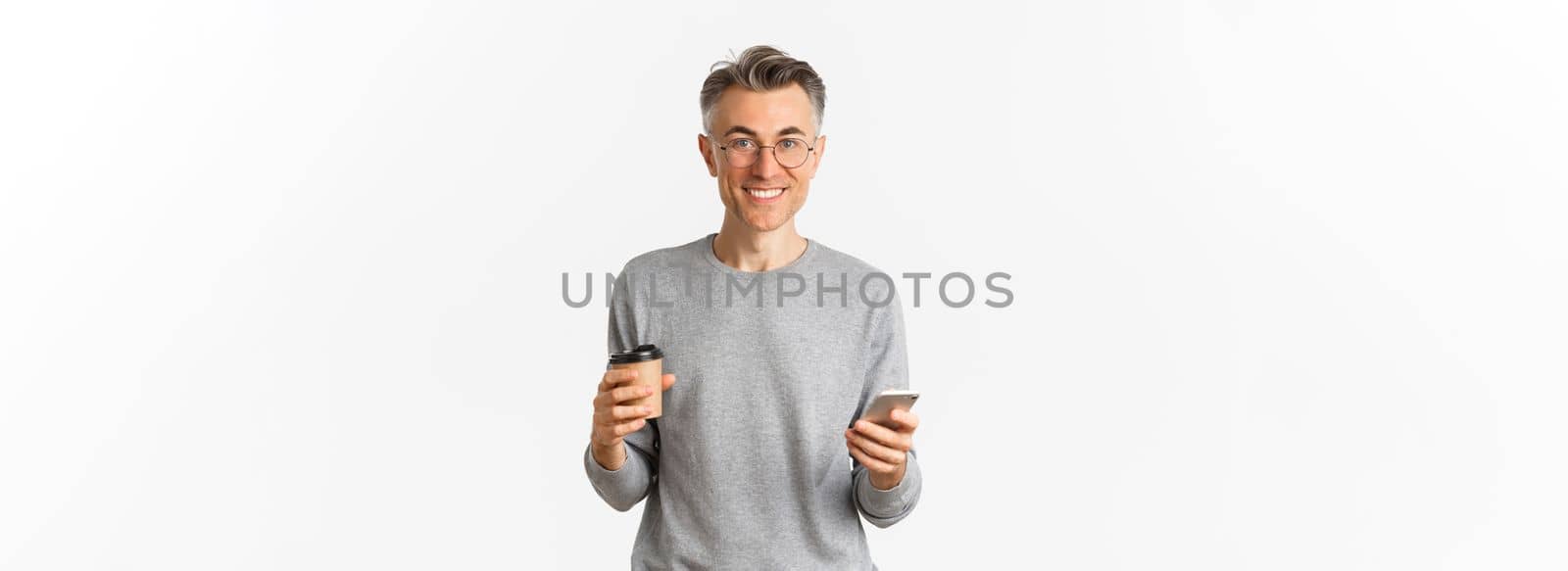 Handsome middle-aged man in grey sweater and glasses, smiling happy, drinking coffee and using mobile phone, standing over white background.