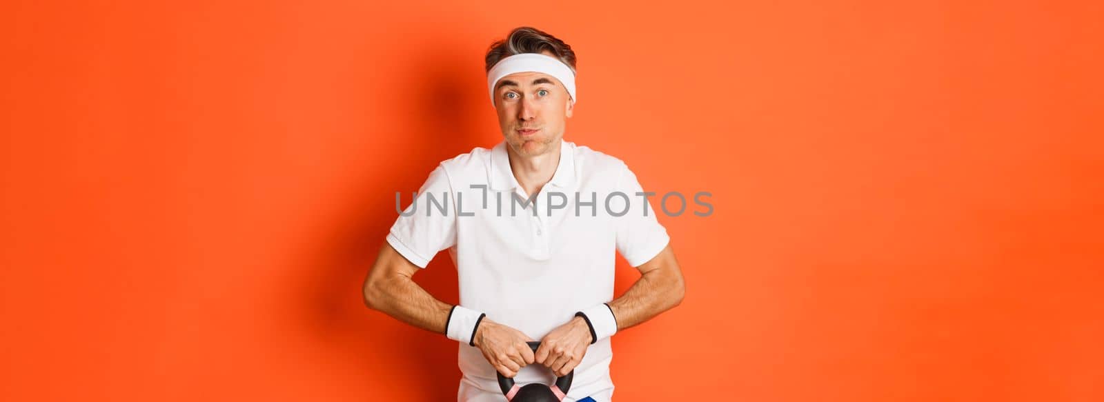 Concept of workout, gym and lifestyle. Image of handsome middle-aged guy doing sport exercises, lifting kettlebell with tensed expression, standing over orange background.