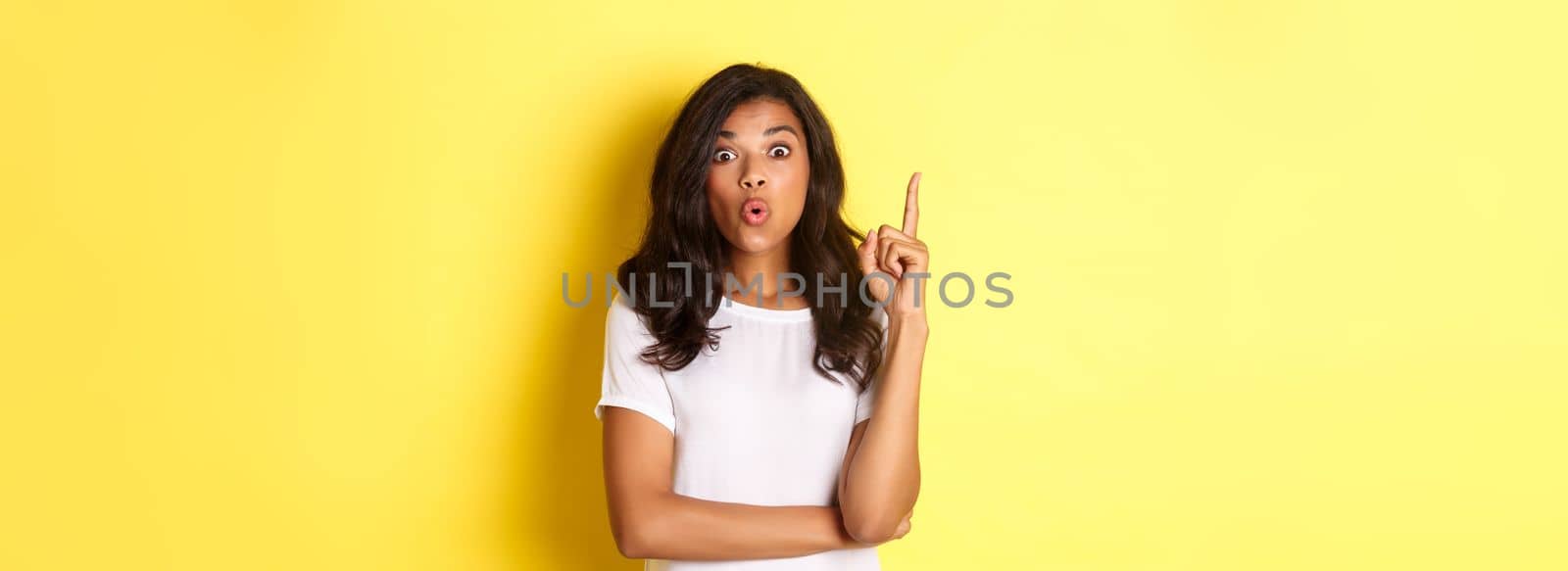 Portrait of smart african-american female student having a solution, suggesting idea, raising finger and looking excited, standing over yellow background.