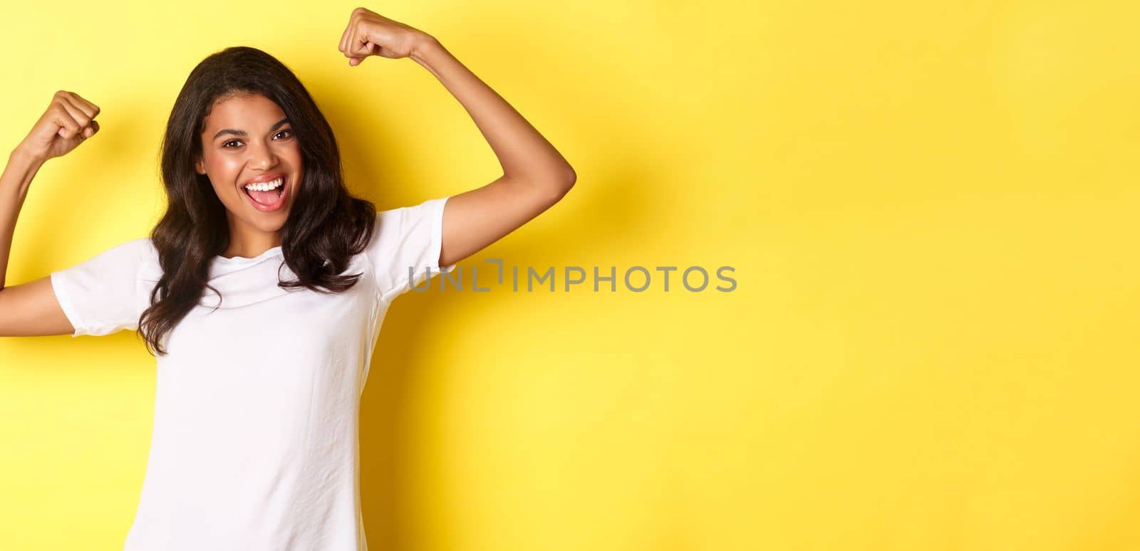 Image of confident and cheerful african-american girl, flexing biceps and smiling, looking strong and happy, standing over yellow background.