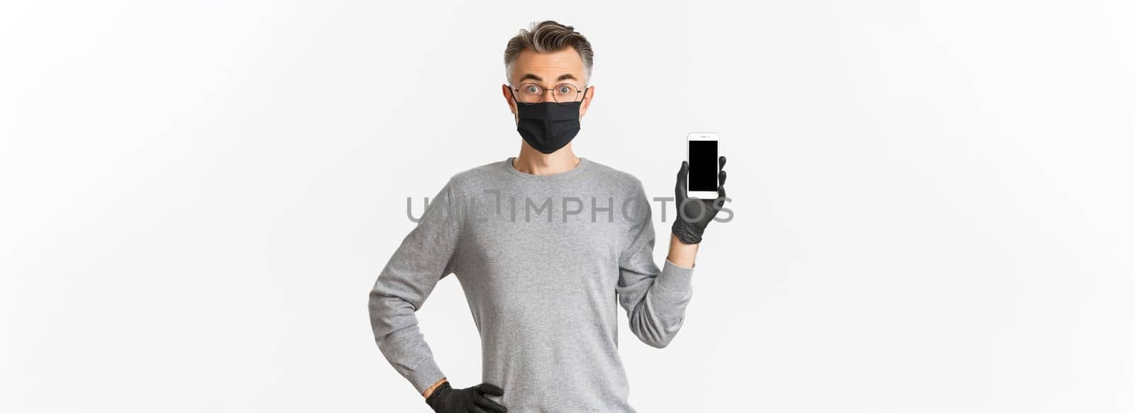 Concept of covid-19, social distancing and lifestyle. Image of surprised middle-aged man found something online, showing smartphone screen and looking amazed, wearing medical mask with gloves.