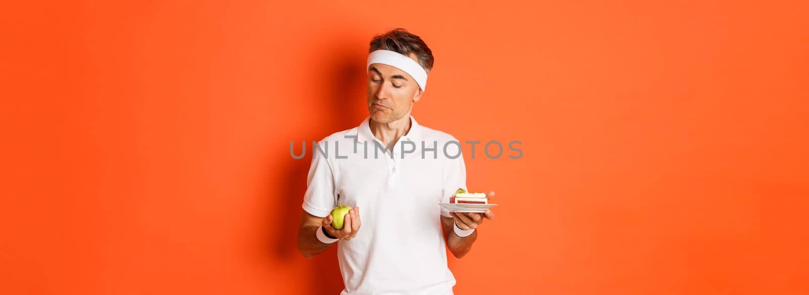 Portrait of handsome middle-aged man in good shape, wearing uniform for training, choosing between apple and cake, standing over orange background.