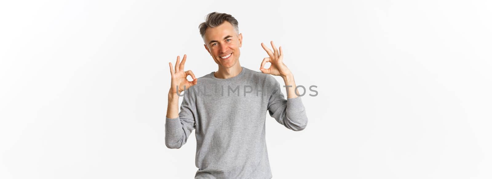 Portrait of successful handsome man in grey sweater, showing okay signs and smiling satisfied, approve something good, recommending product, standing over white background.