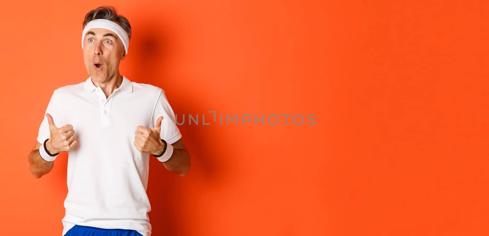 Concept of sport, fitness and lifestyle. Portrait of amazed and excited middle-aged sportsman, looking at upper left corner at something awesome, showing thumbs-up, orange background.