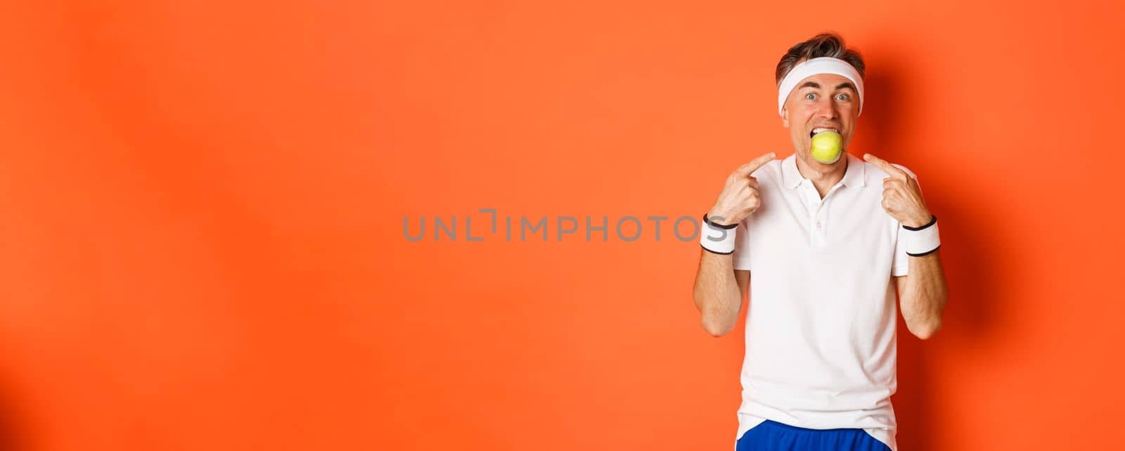 Portrait of funny, handsome middle-aged fitness guy, holding apple with teeth and pointing at it, eating healthy and working out, standing over orange background.