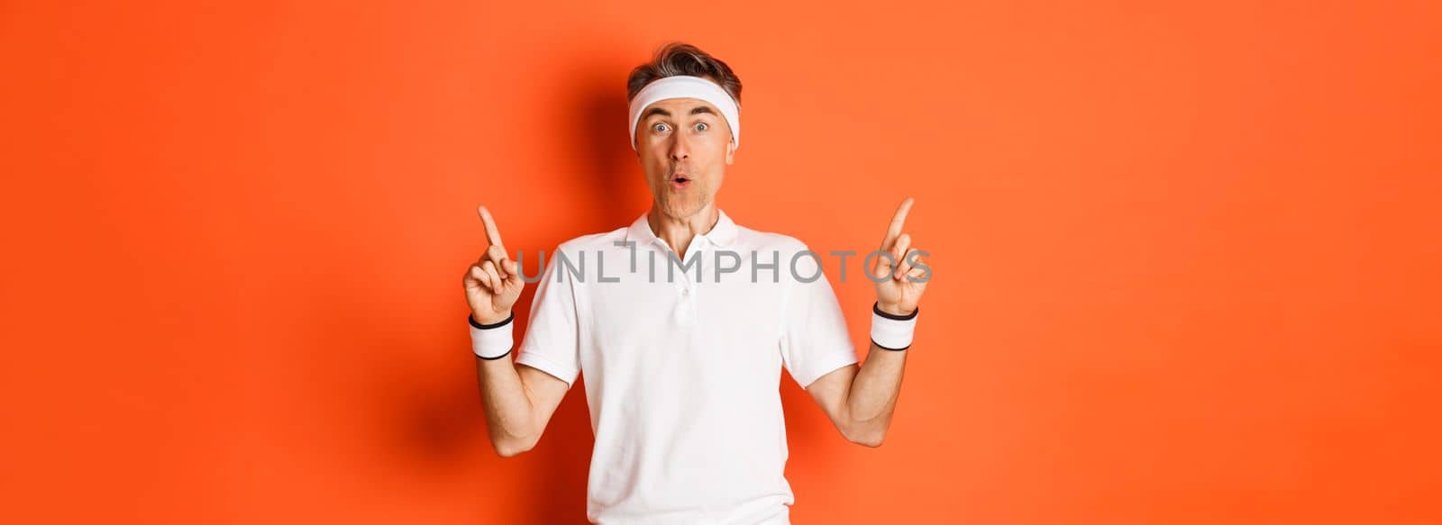 Portrait of handsome middle-aged guy in sports clothing, pointing fingers up, showing promo banner about gym or workout, standing over orange background.