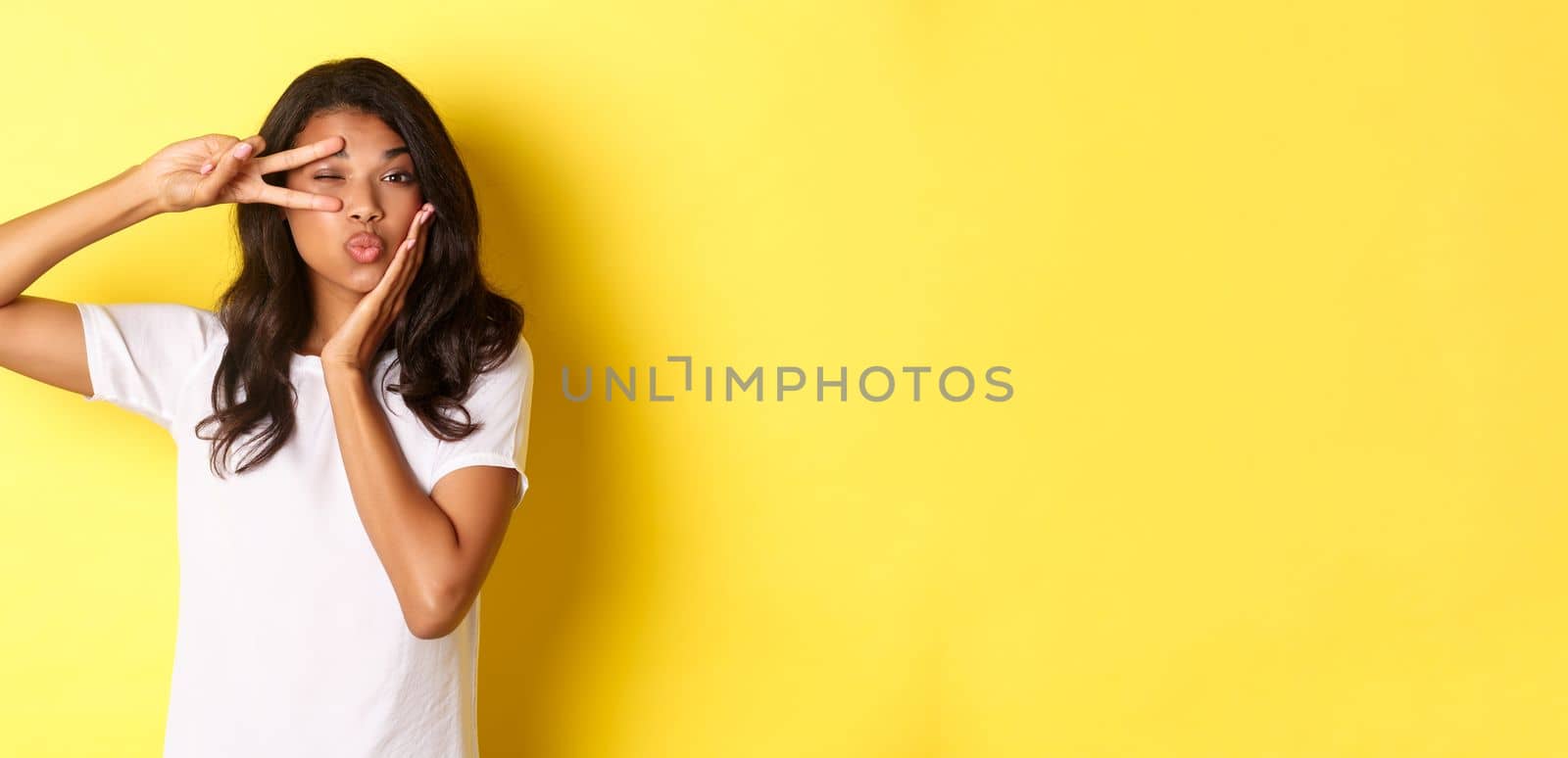 Portrait of beautiful african-american female model, showing peace sign and pouting flirty, standing silly over yellow background.