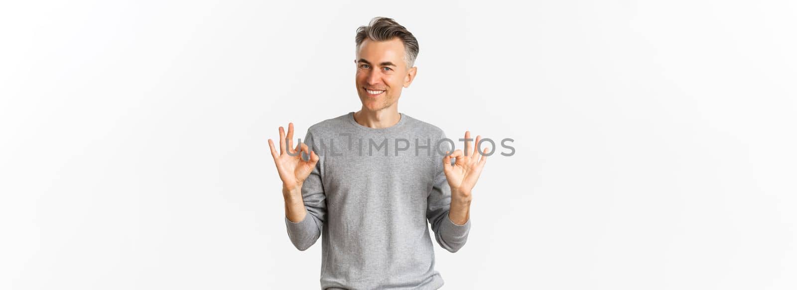 Portrait of handsome middle-aged man, smiling and looking confident while showing okay signs, guarantee something is good, standing over white background.