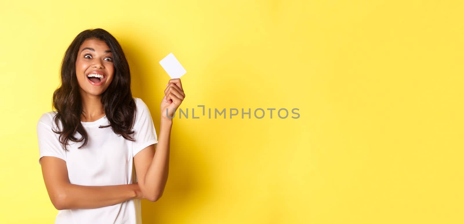 Image of excited african-american woman, showing her credit card and looking amazed at something, going shopping, standing over yellow background.