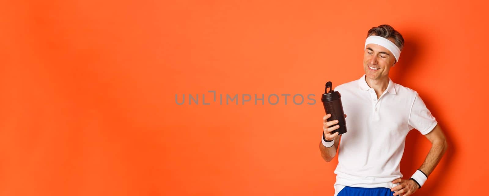 Portrait of pleased middle-aged fitness guy, drinking water and smiling satisfied after productive workout, standing over orange background by Benzoix