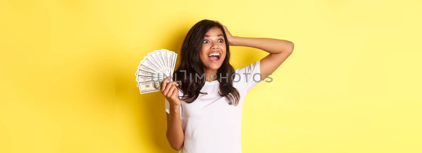 Image of lucky african-american girl, looking excited at upper left corner, holding money, going shopping, standing over yellow background.