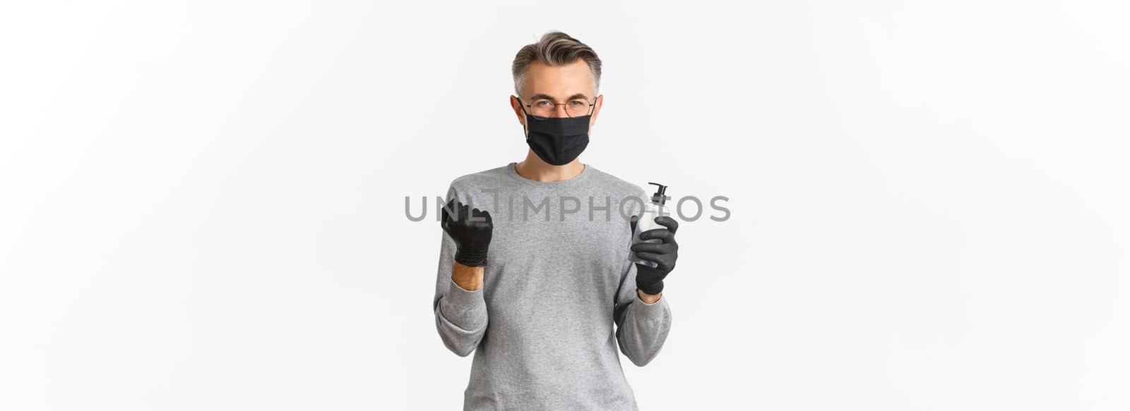Concept of coronavirus, lifestyle and quarantine. Image of cheerful caucasian man in medical mask and gloves, showing hand sanitizer and making fist pump to rejoice, standing over white background.