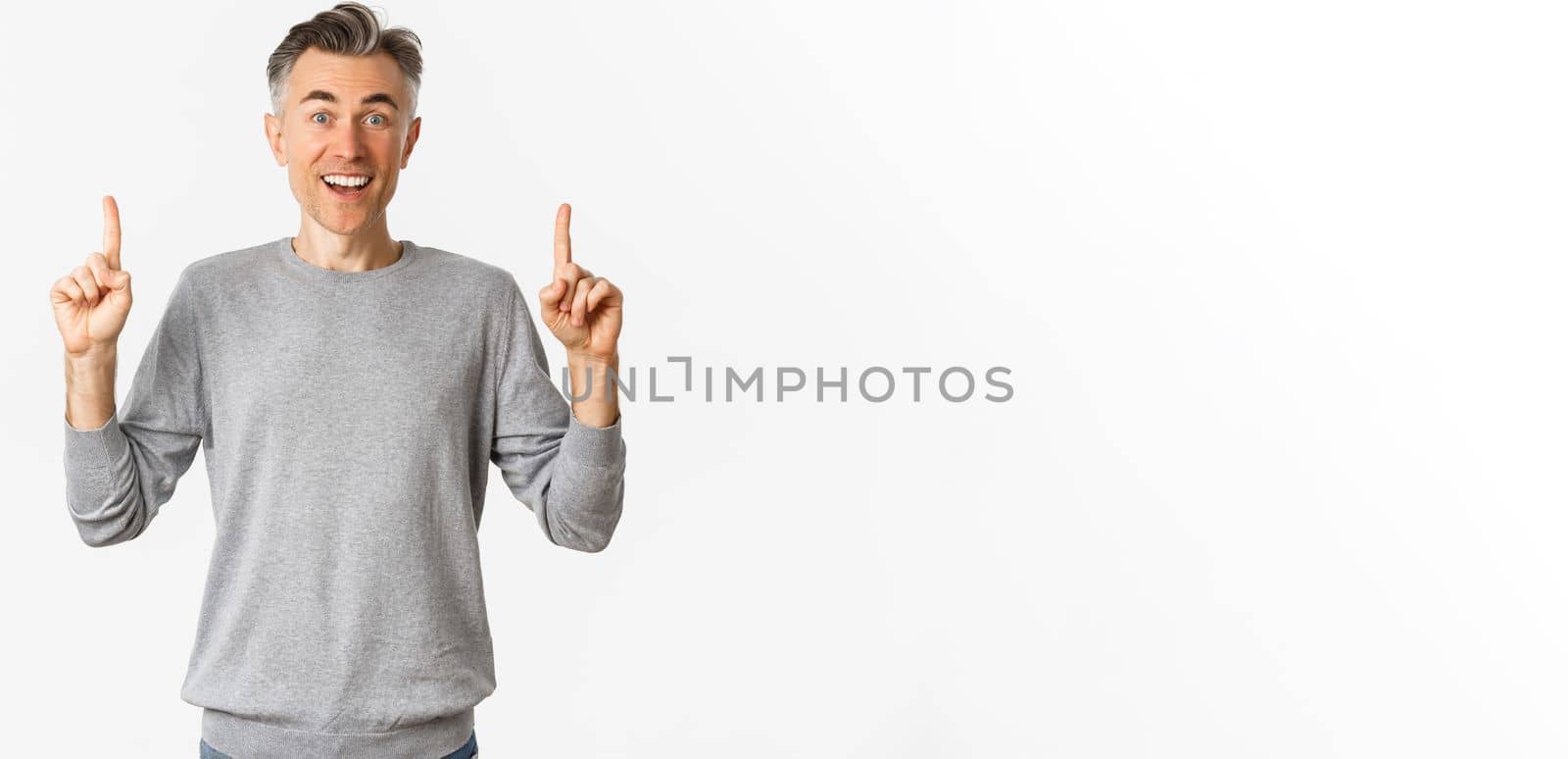 Image of amazed adult guy with grey short hairstyle, showing awesome offer, pointing fingers up at copy space and smiling happy, standing over white background by Benzoix