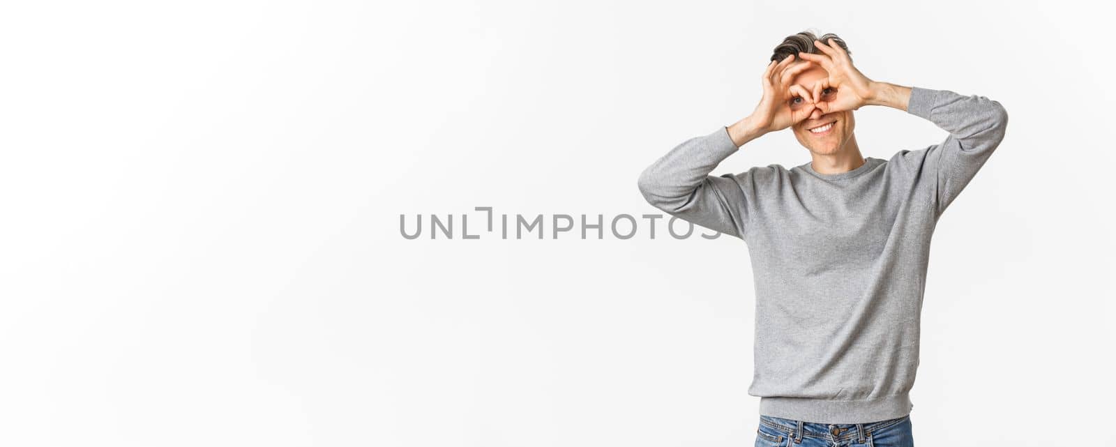 Portrait of funny middle-aged guy making faces, showing hand glasses around eyes as if looking through binoculars, standing over white background.